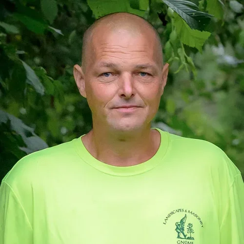 A man with a shaved head standing outdoors among green foliage, wearing a bright yellow T-shirt with a logo on the chest.