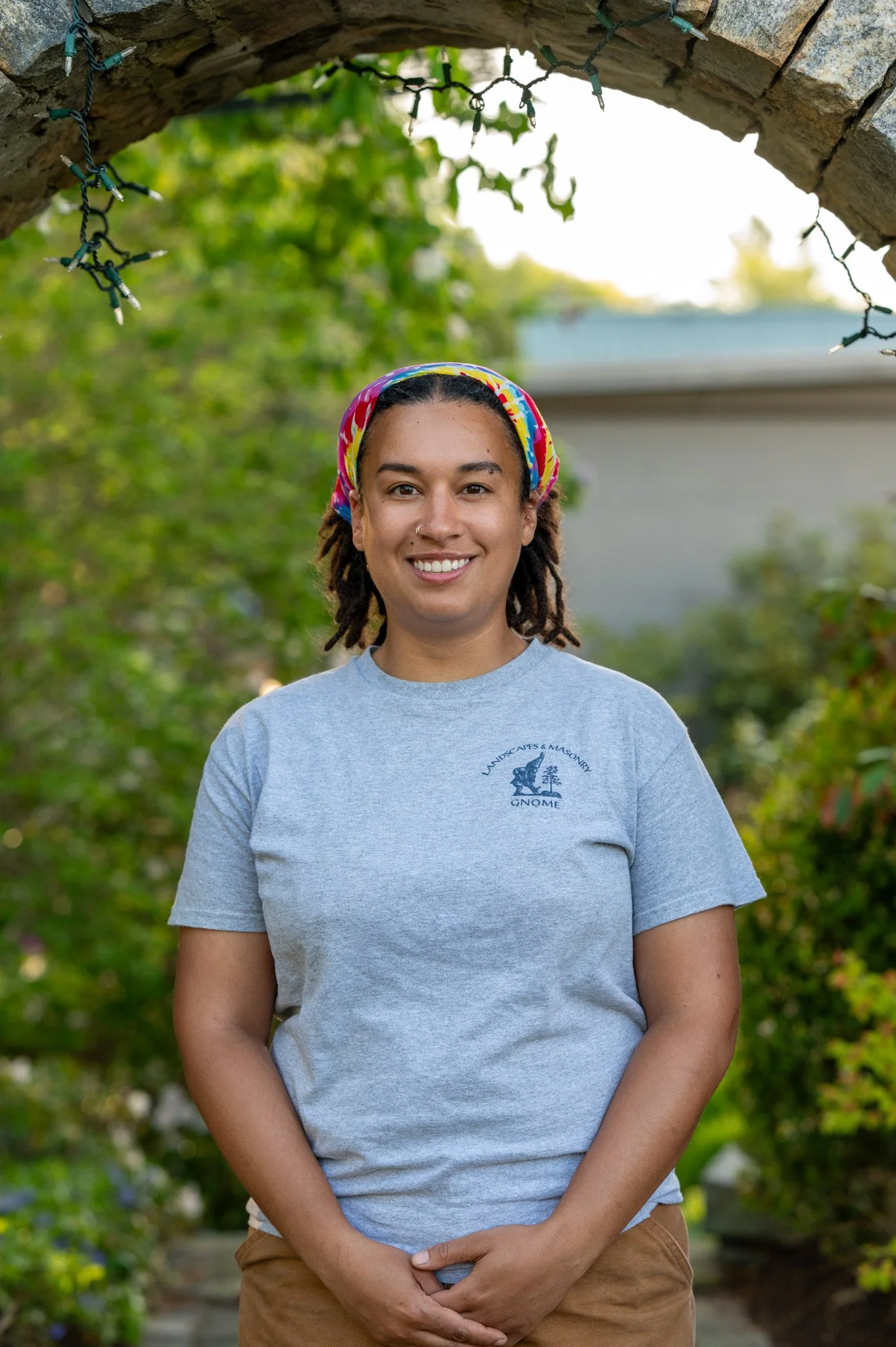 A woman with dreadlocks, wearing a colorful headscarf and a gray T-shirt, standing outdoors under an arch with string lights, smiling at the camera.