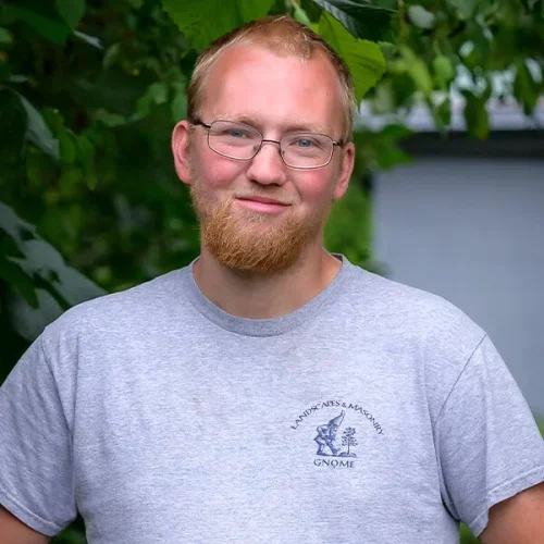 A young man with glasses and a beard smiling outdoors in front of green foliage, wearing a gray T-shirt with a gnome graphic.
