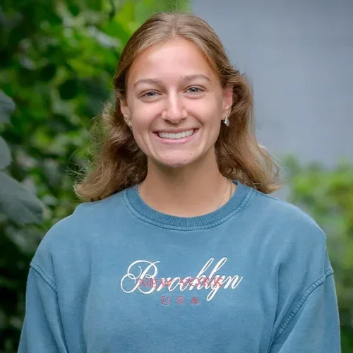 Smiling young woman with long blond hair wearing a blue sweatshirt that says Brooklyn, USA, outdoors with green foliage in the background.
