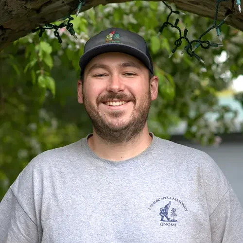 Smiling man outdoors wearing a gray T-shirt and baseball cap, standing under a tree with string lights.