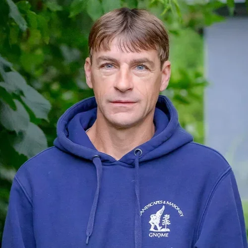A man with short brown hair and blue eyes wearing a blue hoodie with a logo and text on it, standing outdoors with green foliage in the background.