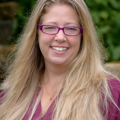 A woman with long blonde hair and purple glasses smiling outdoors.