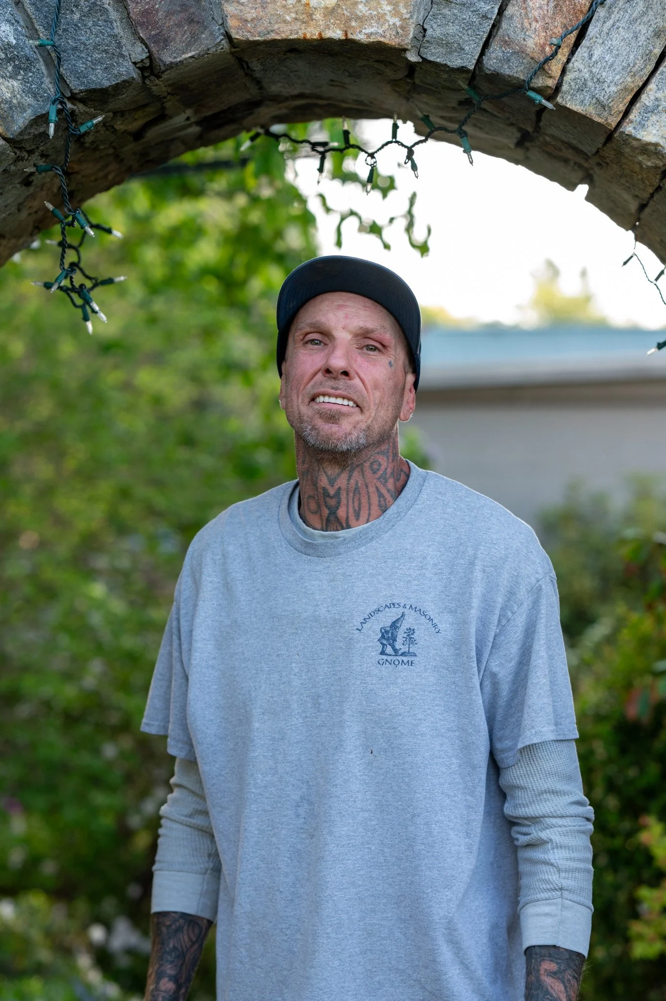 A man with tattoos wearing a grey GNOME t-shirt and a black cap, standing outdoors under an archway decorated with Christmas lights, with trees and a house in the background.