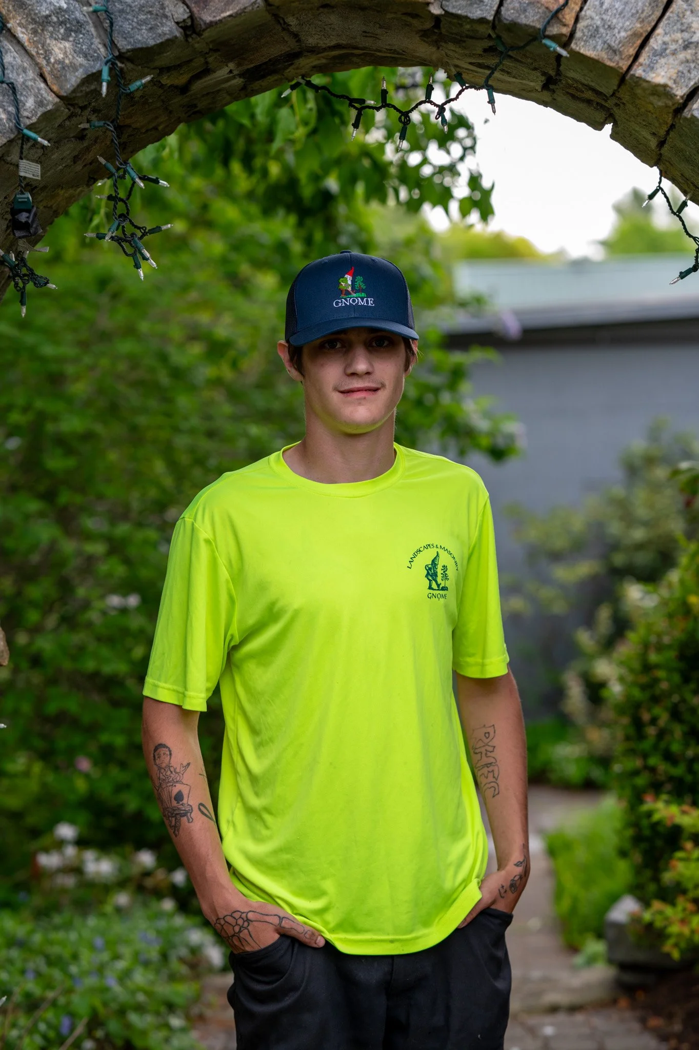A young man wearing a neon yellow T-shirt and a navy baseball cap with the word 'GNOME' embroidered on it. He has tattoos on both arms and is standing outdoors in front of a background with greenery and a building. The photo is taken through an archway decorated with string lights.