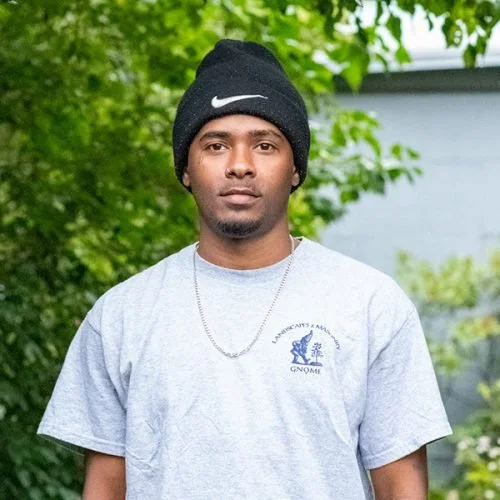 Young man wearing a black Nike beanie and a gray T-shirt with a logo, standing outdoors with green trees in the background.