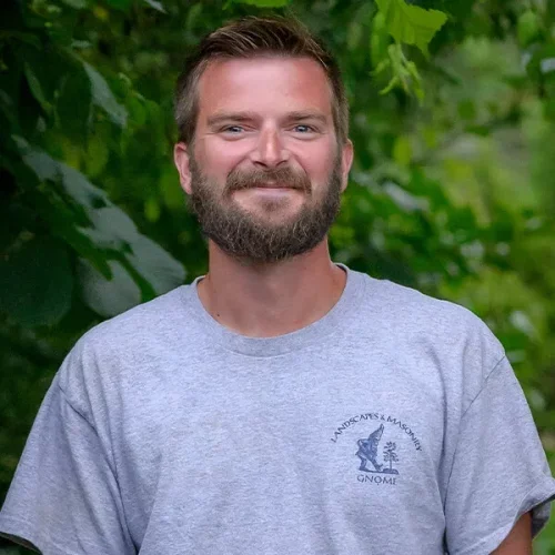 A man with a beard and short hair standing outdoors with green foliage in the background, smiling while wearing a gray t-shirt with a logo.