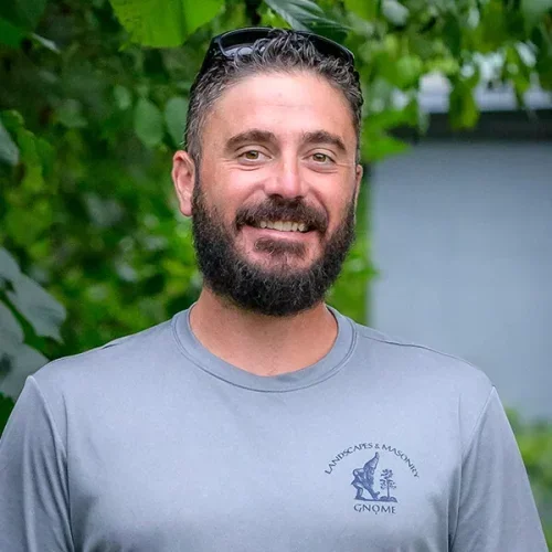 Man with dark hair and beard smiling, wearing sunglasses on his head and a gray T-shirt with a logo.