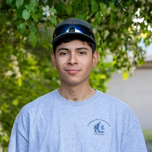 A young man wearing a helmet and sunglasses on his head, standing outdoors in front of green leafy trees, wearing a gray t-shirt with a logo.