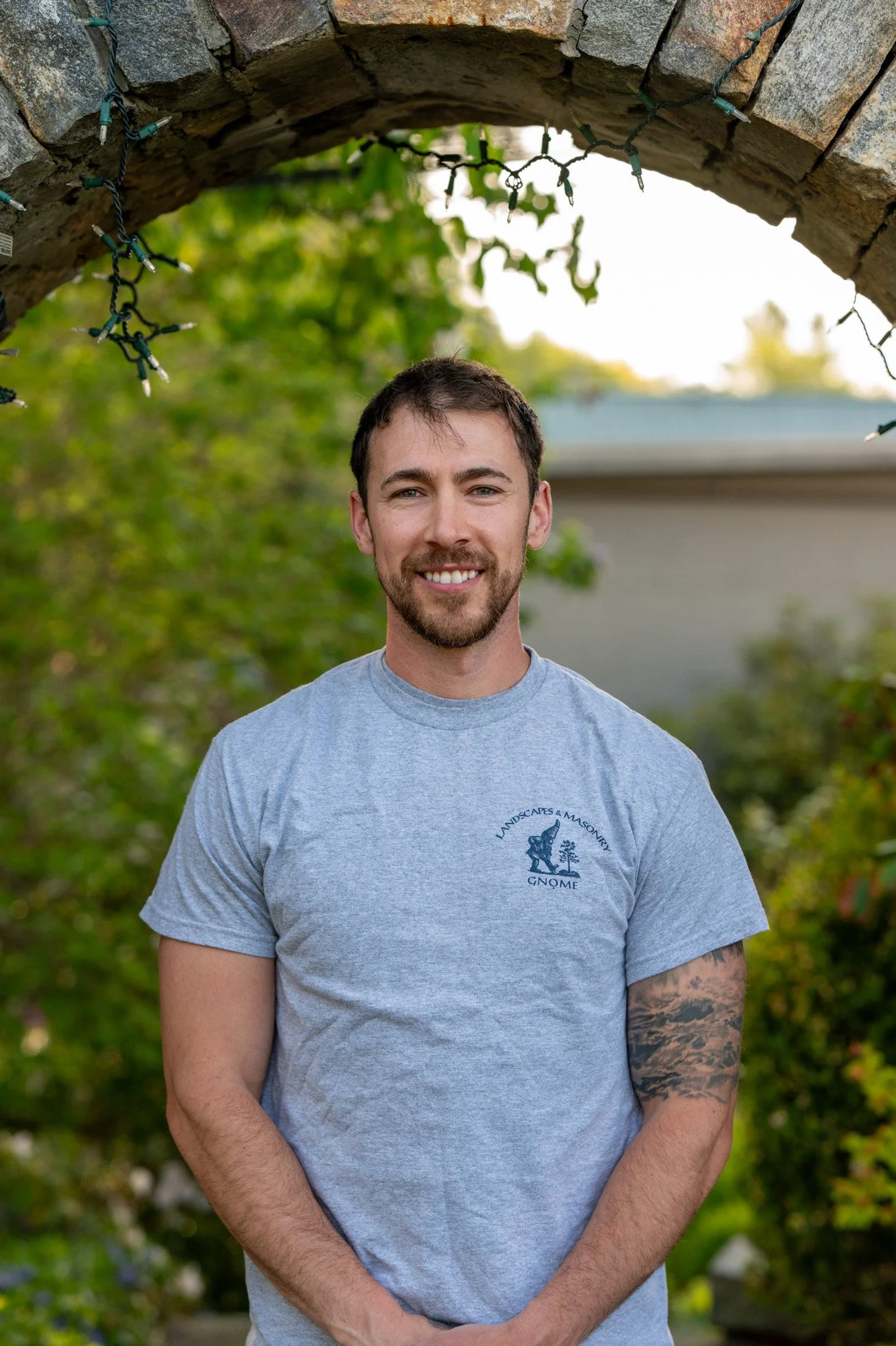 A young man with short dark hair and a beard is smiling and standing outdoors. He is wearing a gray T-shirt with a logo on it. The background includes greenery and a building, and there are old stone and brick elements with stringed Christmas lights hanging above him.