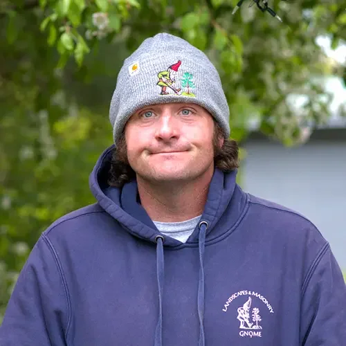 A man with long brown hair wearing a gray beanie with a Peanuts character and a Christmas tree embroidery, smiling outdoors with green trees in the background. He is wearing a navy blue hoodie with a logo on the chest.