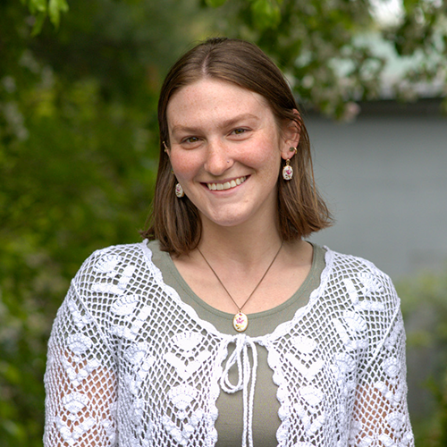 Young woman smiling outdoors, wearing a white crochet cardigan, earrings, and necklace.