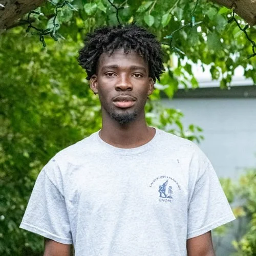 Young man with curly hair and a beard wearing a gray t-shirt standing outdoors with green foliage in the background.