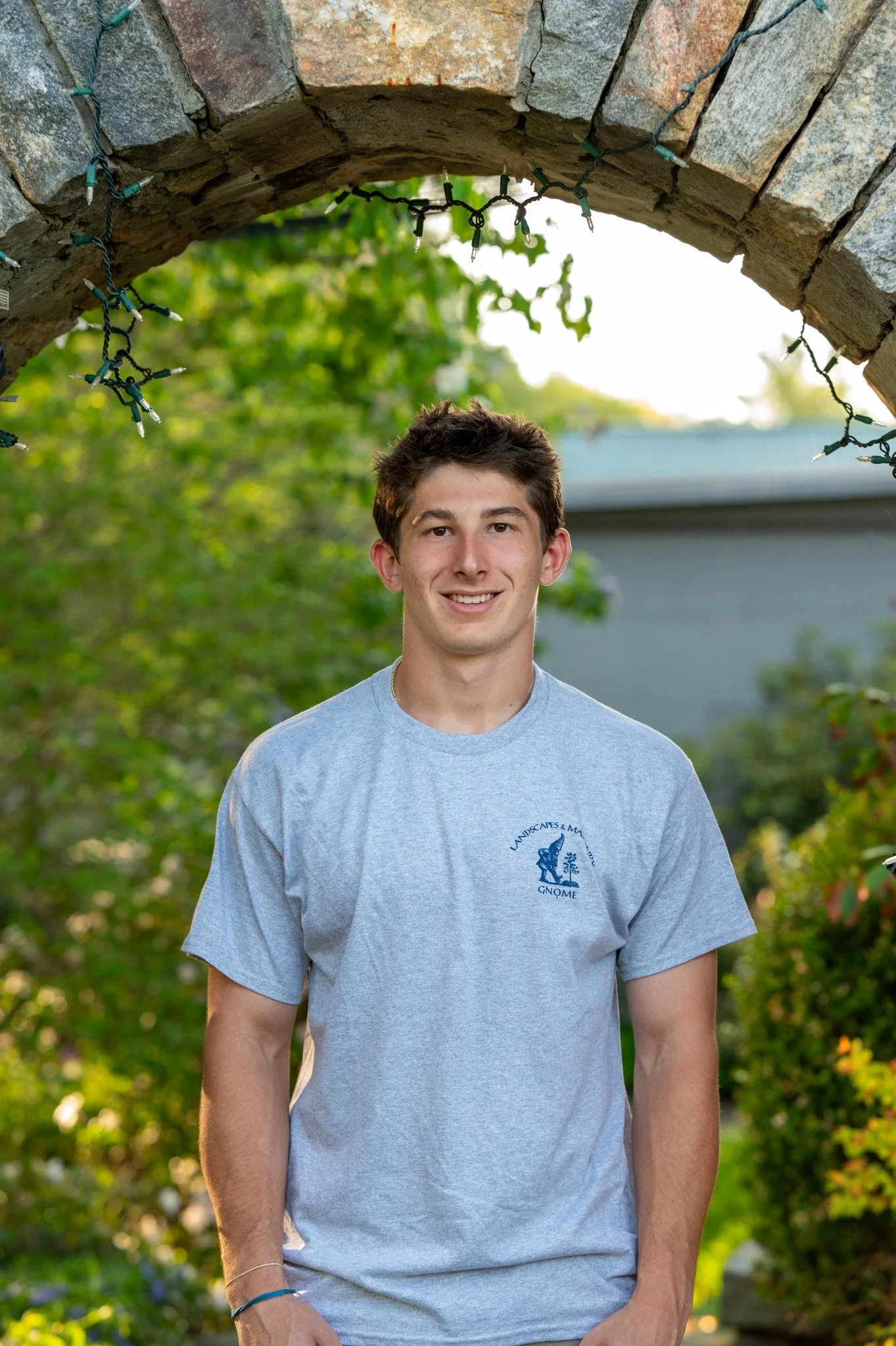 A young man with short dark hair smiling outdoors under a stone archway decorated with string lights. He is wearing a light gray T-shirt with a blue graphic and text. The background features green trees and a blue building.
