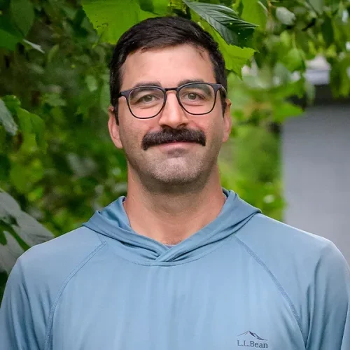 A man with glasses and a mustache wearing a light blue LL Bean hoodie standing outdoors surrounded by green leaves.