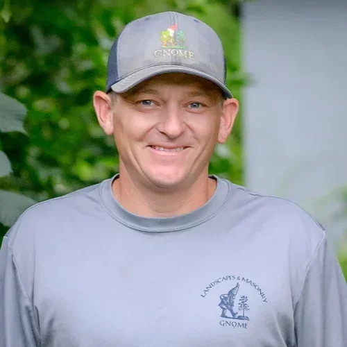 Man smiling outdoors wearing a gray Gnome landscaping and masonry shirt and a gray Gnome cap, with greenery in the background.