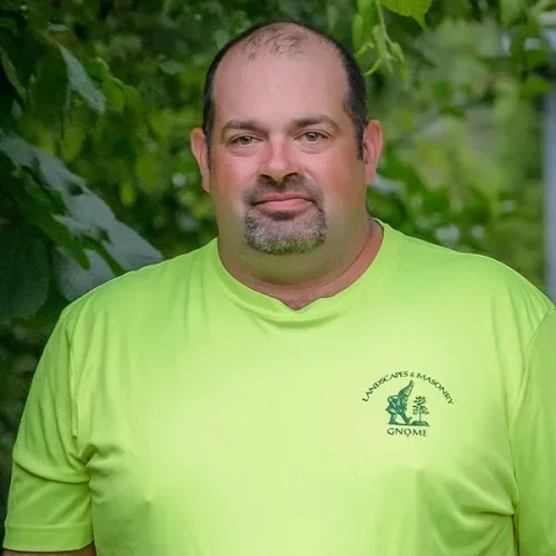 A man with a beard and short hair, wearing a bright green T-shirt, standing outdoors with greenery in the background.
