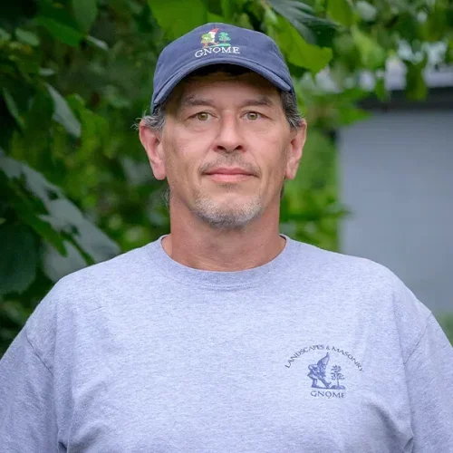 A man wearing a Gnome hat and a gray t-shirt with a gnome logo, standing outdoors with green foliage in the background.