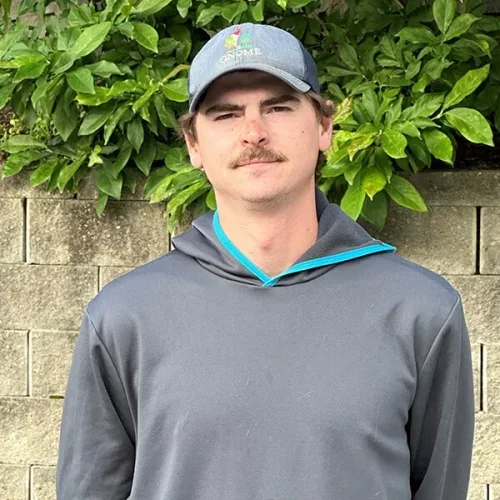 Young man with a mustache wearing a gray hoodie and a gray cap with a logo, standing outdoors in front of green foliage and a stone wall.