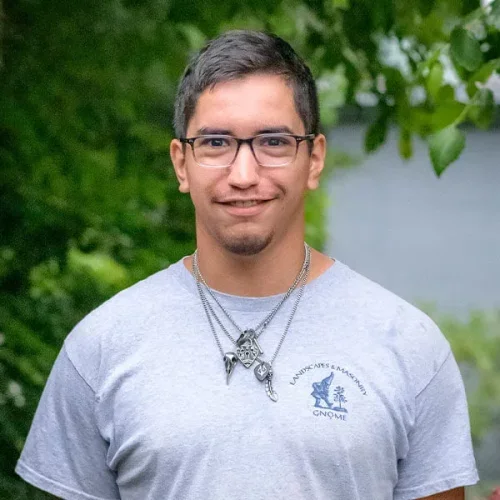 A man with glasses and short dark hair smiling outdoors, wearing a light gray T-shirt with a logo on the left side and multiple necklaces, with green trees and a body of water in the background.