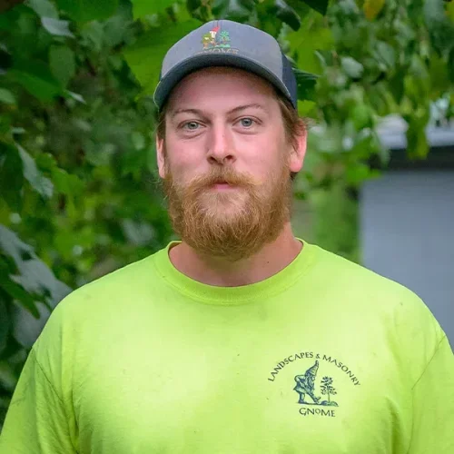 A man with a beard and mustache wearing a gray cap and neon green T-shirt standing outdoors with greenery in the background.