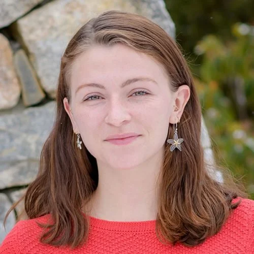 A young woman with long brown hair, wearing a red top and star-shaped earrings, standing outdoors near a stone wall with green foliage in the background.