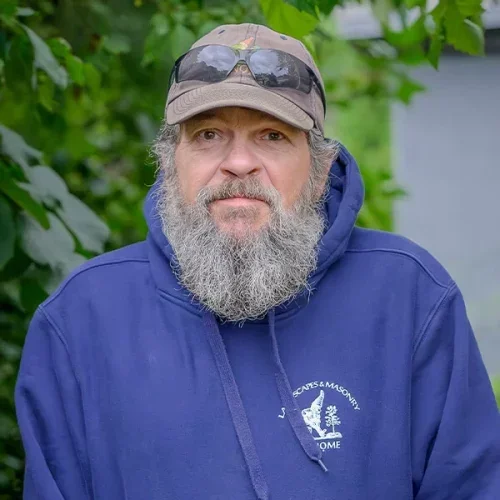 A bearded man wearing a khaki baseball cap, sunglasses on top, and a blue hoodie with a logo on it, standing outdoors with green foliage in the background.