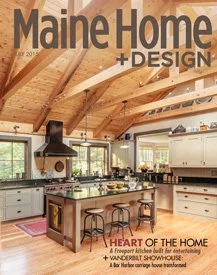 Cover of Maine Home + Design magazine featuring a modern kitchen with wooden ceiling beams, white cabinets, large windows, and a kitchen island with barstools