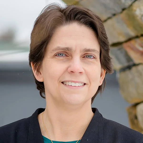 Headshot of a woman with short brown hair and blue eyes, smiling outdoors with rocks in the background.