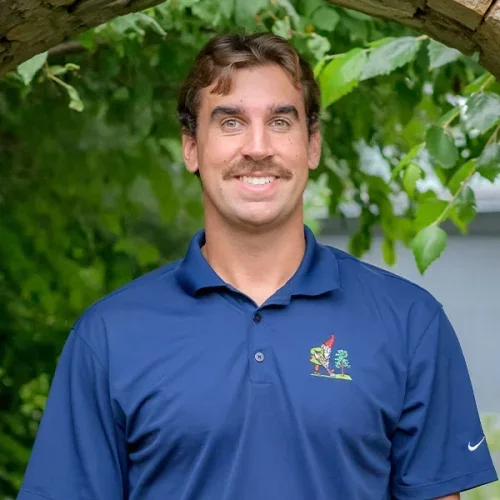 A man with light brown hair and a mustache, wearing a blue polo shirt with a colorful logo, standing outdoors with green foliage in the background.