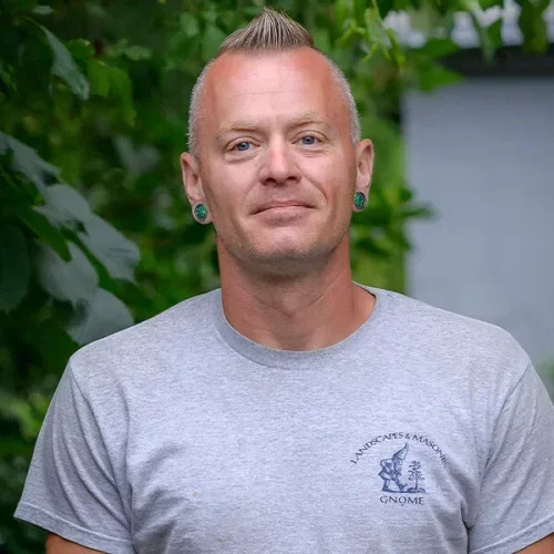 A man with short blond hair, wearing a light gray T-shirt and green earrings, standing outdoors with green foliage in the background.