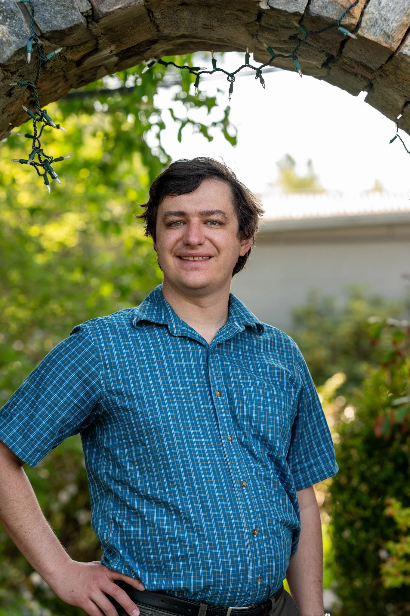 Young man in a blue checkered shirt standing outdoors under a brick arch with string lights, with greenery and trees in the background.