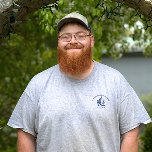 Smiling man with glasses, red beard, gray shirt, and cap outdoors under a tree.