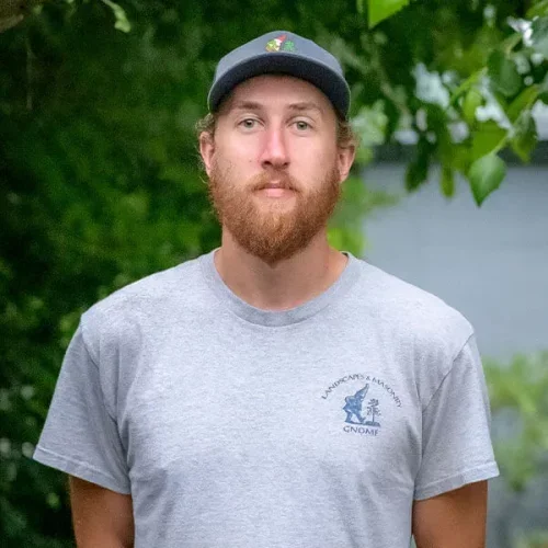 A young man with a red beard and mustache wearing a gray T-shirt and a gray baseball cap, standing outdoors with green trees and a body of water in the background.