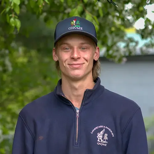 Young man wearing a navy blue jacket and a black cap with floral design, outdoors with trees in the background.