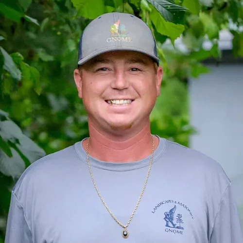 A man smiling outdoors wearing a gray Gnome baseball cap and a gray T-shirt with a logo for landscaping and masonry, with a chain necklace, standing in front of green foliage.