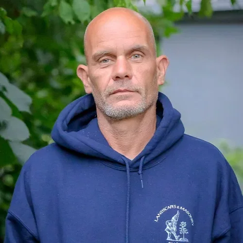 A middle-aged man with a shaved head and a trimmed beard, wearing a navy blue hoodie with a lakes and mountains logo, standing outdoors with greenery in the background.