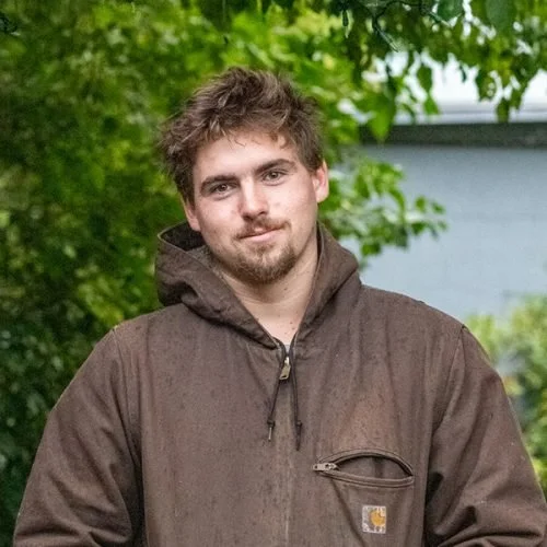 A young man with light brown hair and a beard wearing a brown jacket, standing outdoors with green trees and a house in the background.