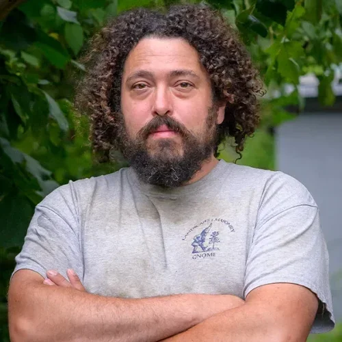 A man with curly hair and a beard standing outdoors with his arms crossed, wearing a gray T-shirt, greenery in the background.