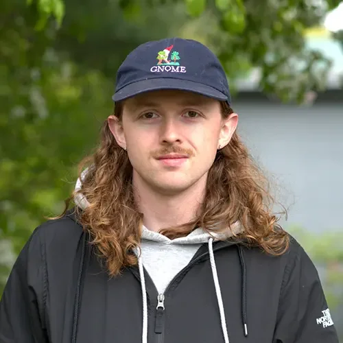 A young man with long, curly hair wearing a navy baseball cap with a Gnome logo, a light gray hoodie, and a black North Face jacket, outdoors with green trees in the background.