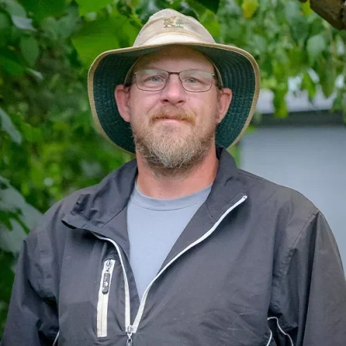 A man with glasses and a beard, wearing a wide-brimmed hat and a gray jacket, standing outdoors with green foliage in the background.