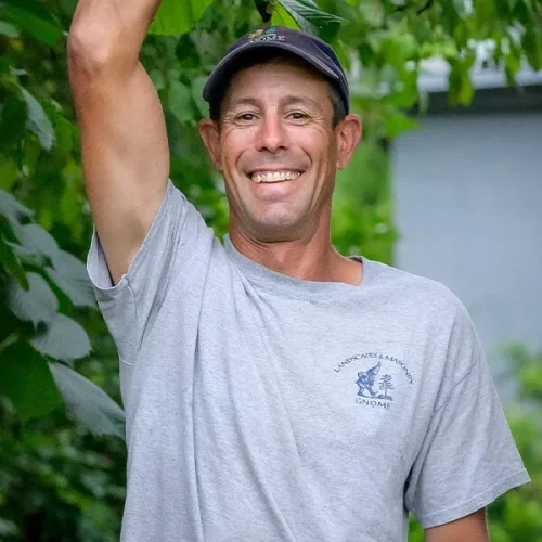 A smiling man wearing a gray t-shirt with a logo, a cap, and raising his arm with greenery in the background.