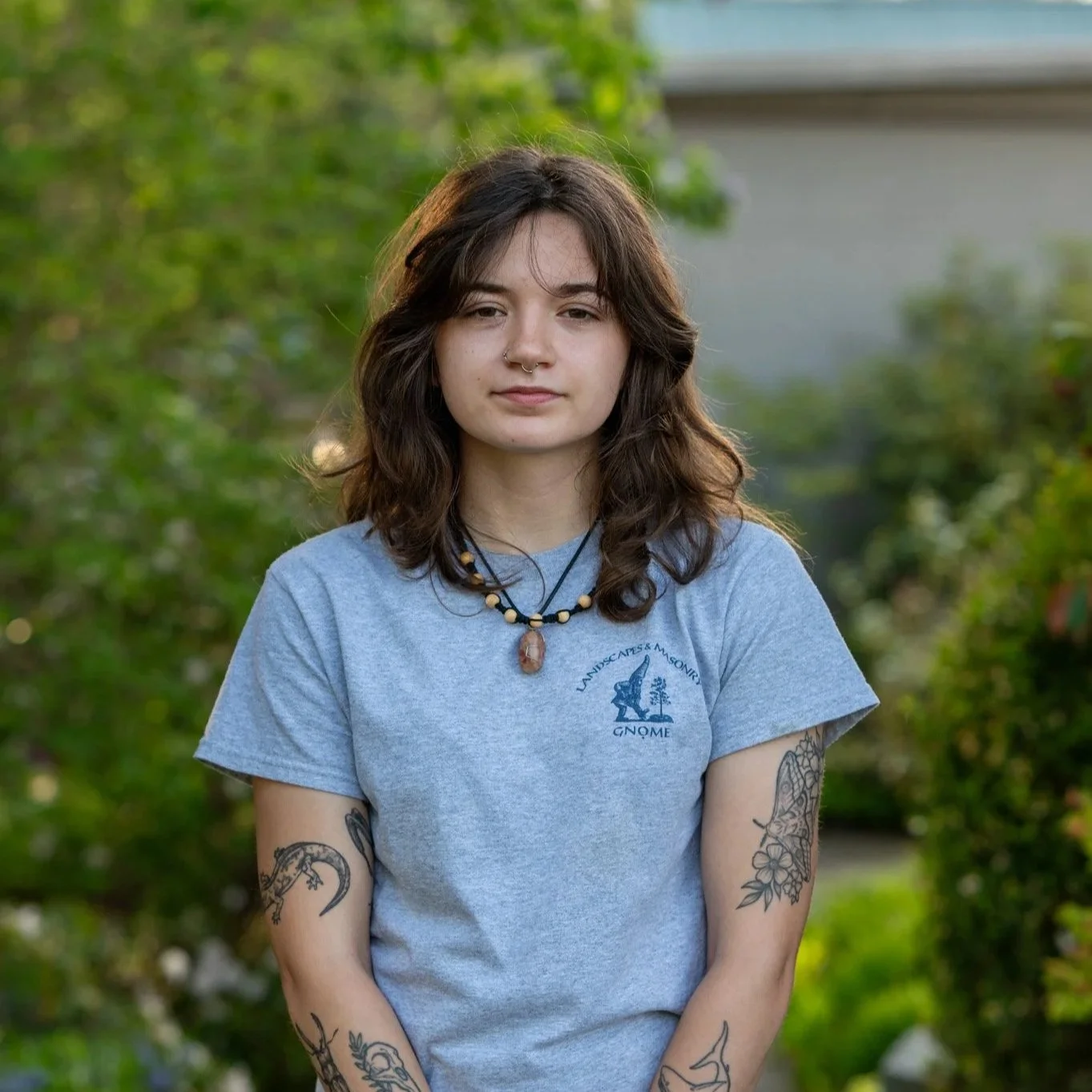 Young woman with shoulder-length wavy brown hair, wearing a grey T-shirt with a logo, and multiple tattoos on her arms, standing outdoors with green foliage in the background.