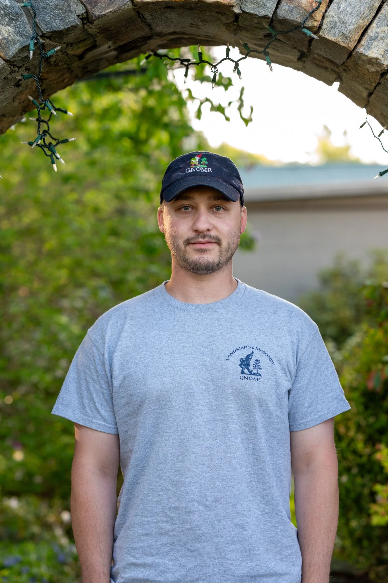 A portrait of a young man with a beard, wearing a black cap and a gray T-shirt, standing outdoors with trees and a house in the background. The cap and T-shirt both have a logo with the text "GNOME" and an illustration of a gnome with a tree.