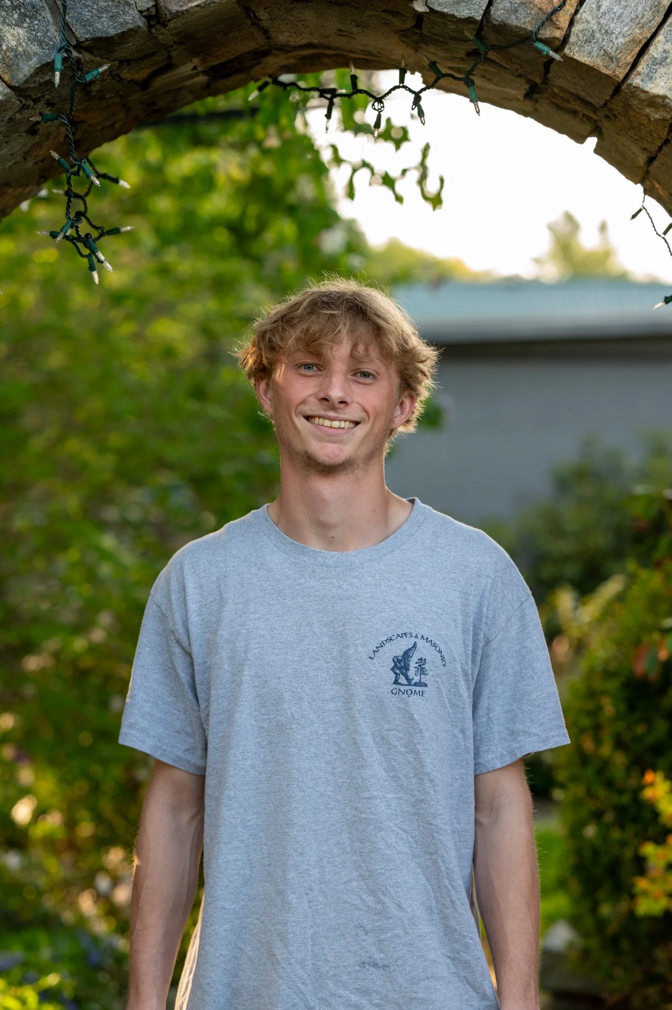 A smiling young man with wavy blonde hair standing outdoors under an archway of a stone structure, with string lights hanging above and greenery in the background.