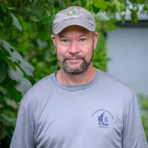A man wearing a gray GNOME cap and gray shirt with a logo, standing outdoors in front of green foliage.