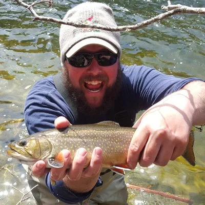 Man with a beard, wearing a baseball cap and sunglasses, holding a large fish near a river.