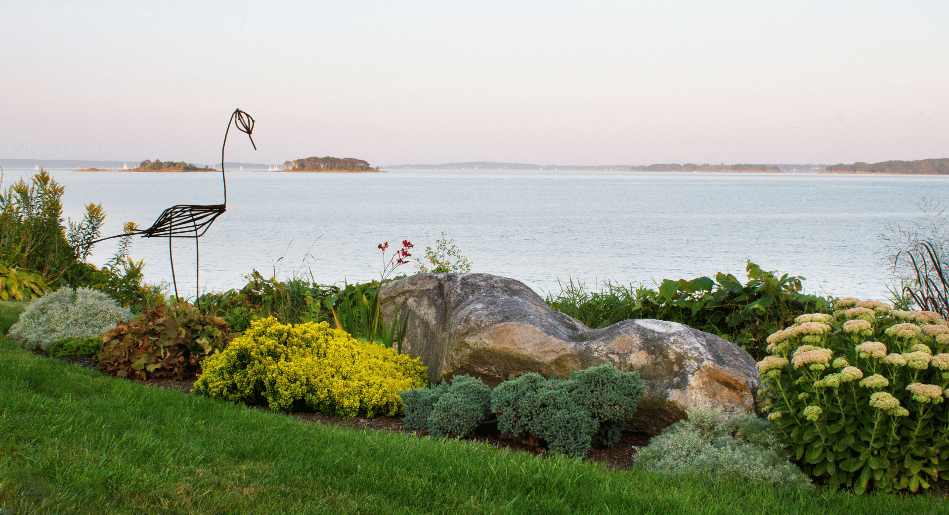 A landscaped garden with green grass, colorful bushes, and flowers, featuring a wire sculpture of a bird near a large rock, overlooking a serene body of water with small islands and sailboats in the distance.