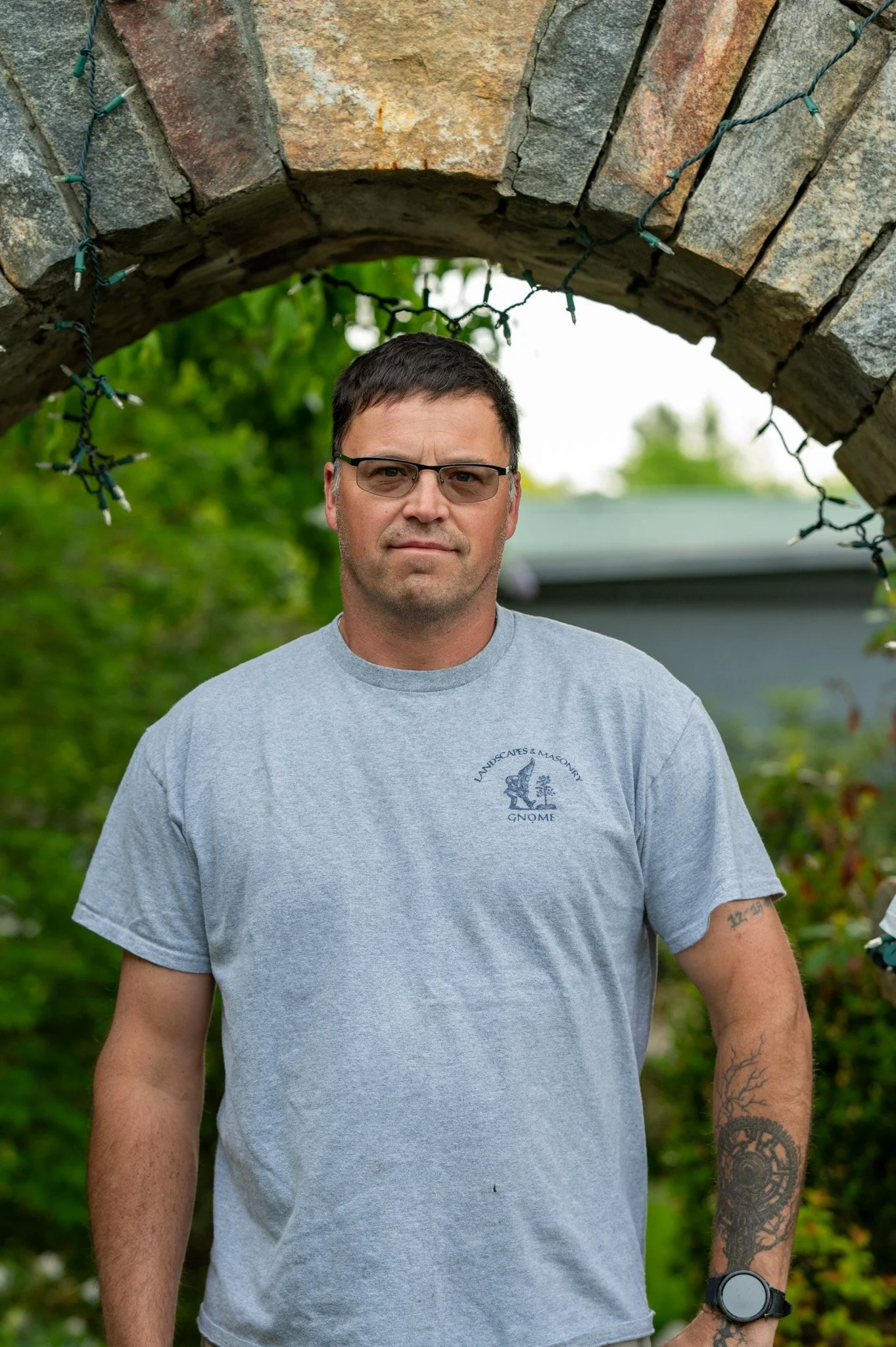 A man with glasses and tattoos on his arms standing outdoors under a brick archway decorated with string lights. He is wearing a gray T-shirt with a logo on the chest that says 'Landscapes & Moonrncy Gnome'.