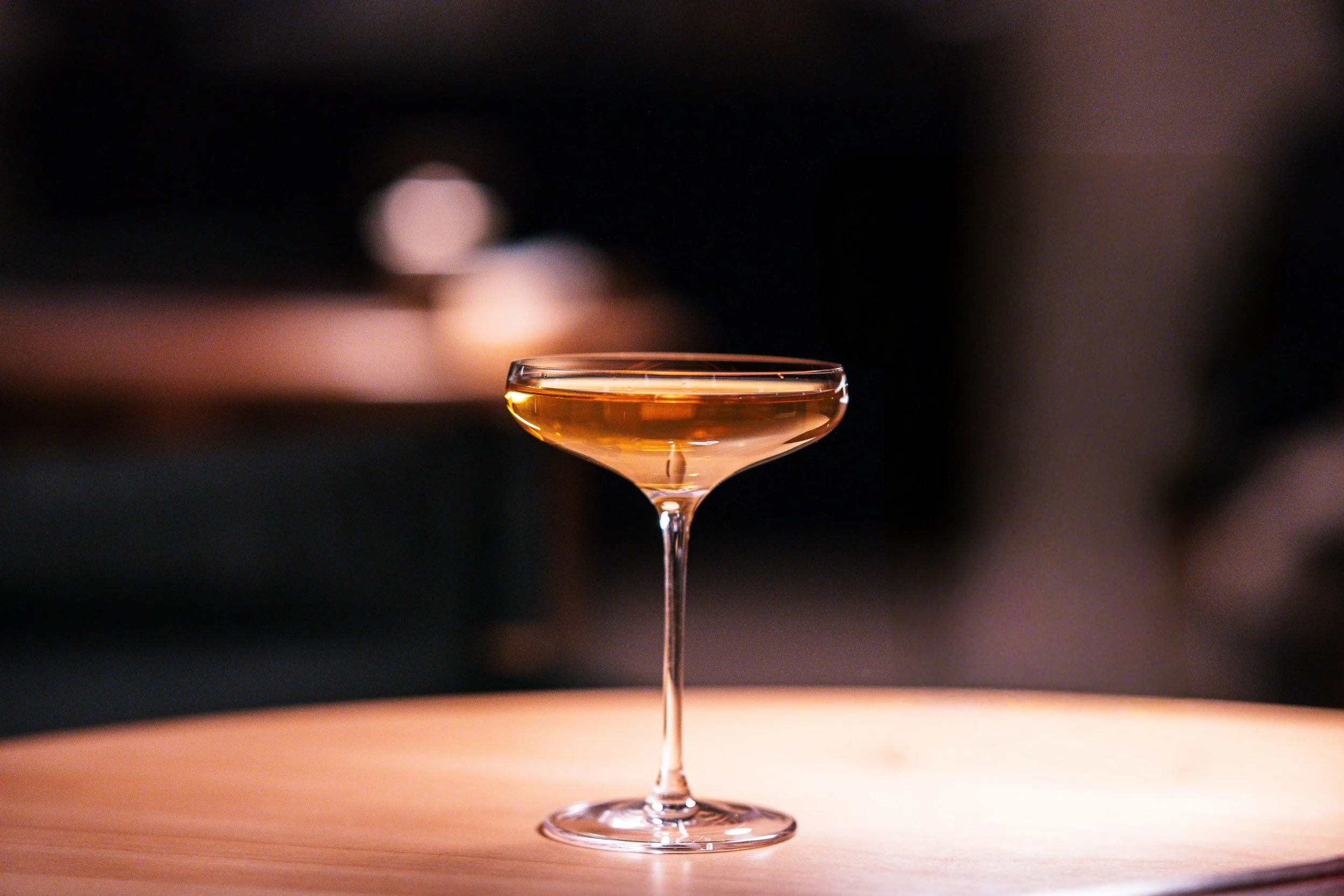 A glass coupe filled with an amber-colored cocktail on a wooden surface with a dark, blurred background.